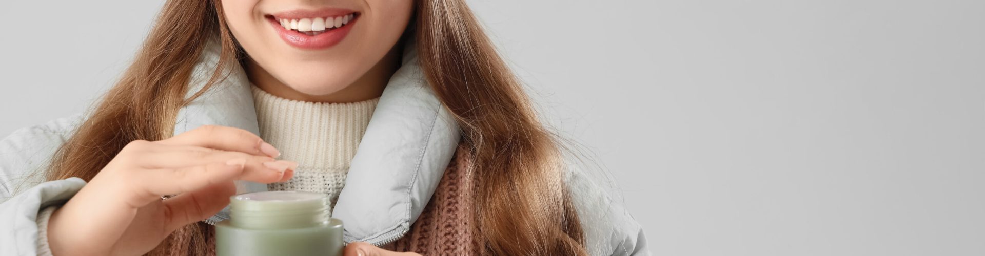 Happy woman in winter clothes and with cosmetic cream on grey background